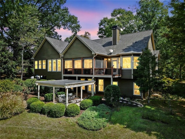 back of property at dusk with a patio, roof with shingles, a pergola, a chimney, and a lawn