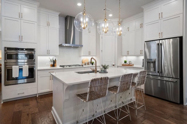 kitchen with stainless steel appliances, a kitchen island with sink, pendant lighting, and wall chimney exhaust hood