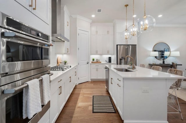 kitchen featuring appliances with stainless steel finishes, white cabinetry, an island with sink, sink, and wall chimney range hood