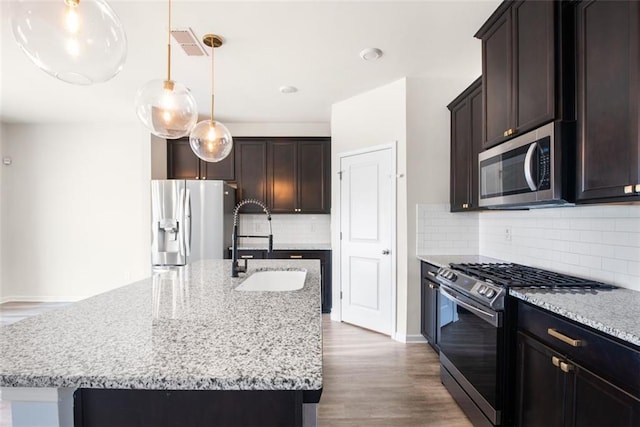 kitchen featuring a kitchen island with sink, stainless steel appliances, hanging light fixtures, light stone counters, and decorative backsplash