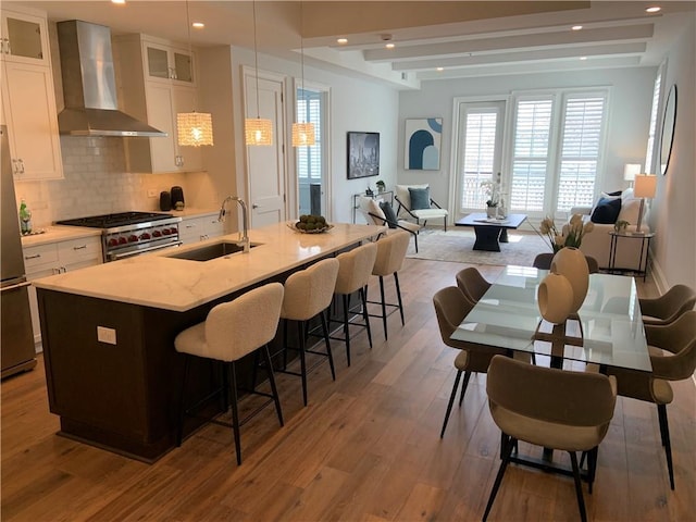 kitchen with sink, white cabinetry, wall chimney range hood, stainless steel appliances, and an island with sink