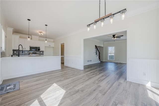 unfurnished living room with visible vents, a wainscoted wall, light wood-style flooring, stairs, and crown molding