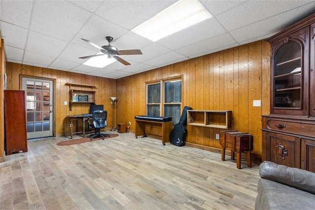 home office with a ceiling fan, light wood-type flooring, a drop ceiling, and wood walls