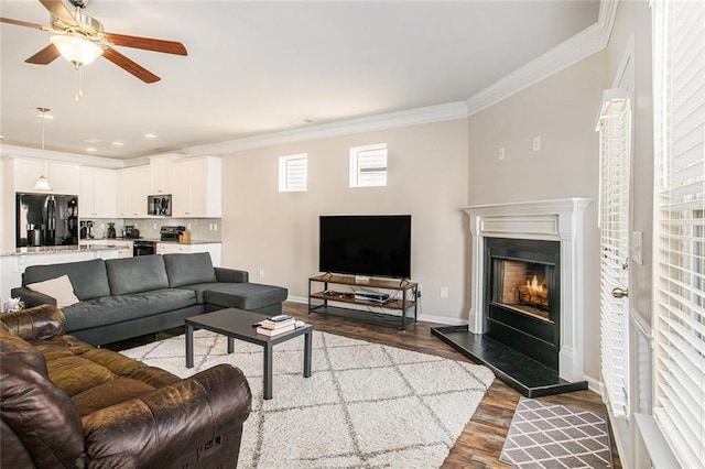 living room with ceiling fan, wood finished floors, baseboards, a lit fireplace, and crown molding