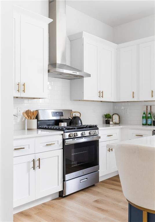 kitchen with wall chimney range hood, white cabinetry, backsplash, stainless steel gas range oven, and light hardwood / wood-style floors