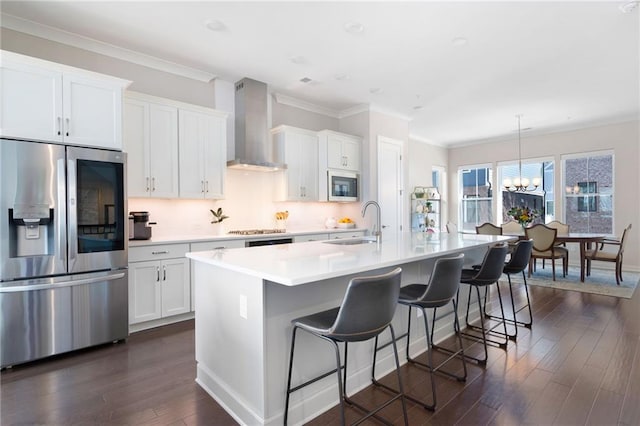 kitchen featuring a kitchen island with sink, dark wood-style flooring, a sink, stainless steel appliances, and wall chimney range hood