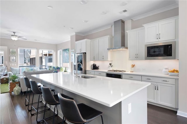 kitchen featuring an island with sink, a sink, stainless steel appliances, a kitchen bar, and wall chimney range hood