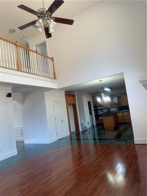 unfurnished living room with crown molding, a towering ceiling, ceiling fan with notable chandelier, baseboards, and dark wood-type flooring
