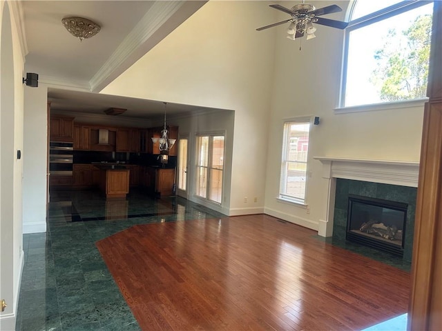 unfurnished living room featuring a ceiling fan, dark wood-style flooring, a tiled fireplace, baseboards, and ornamental molding