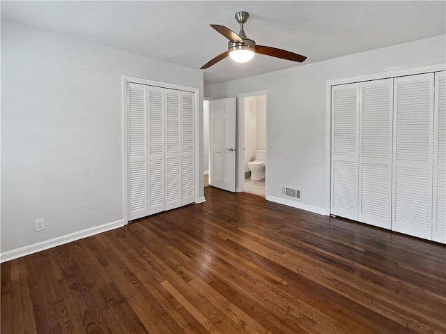 unfurnished bedroom with ceiling fan, ensuite bath, dark hardwood / wood-style floors, and two closets