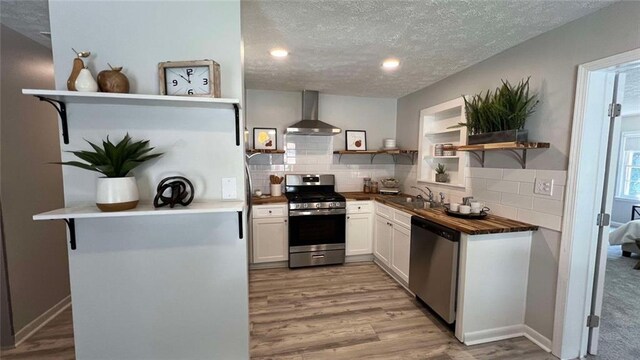 kitchen with light wood-type flooring, a textured ceiling, white cabinets, wall chimney range hood, and stainless steel appliances