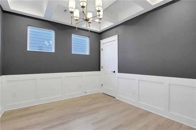 unfurnished dining area with light wood-style flooring, a wainscoted wall, an inviting chandelier, coffered ceiling, and a decorative wall