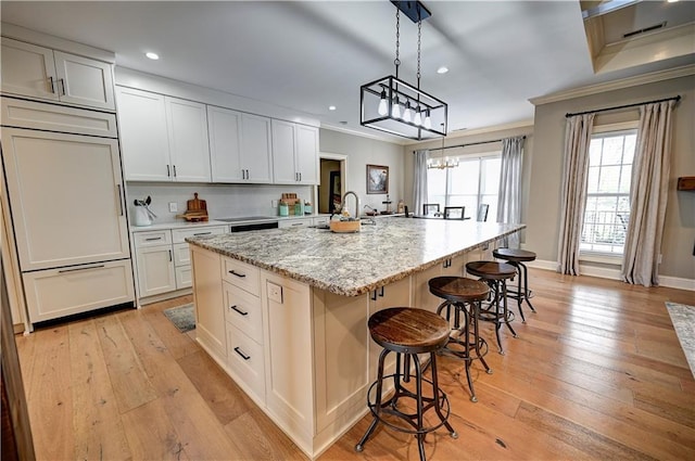 kitchen with a center island with sink, hanging light fixtures, light stone counters, white cabinets, and crown molding