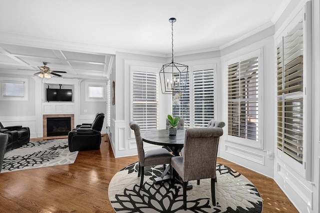 dining area with a tile fireplace, dark hardwood / wood-style floors, ceiling fan with notable chandelier, coffered ceiling, and crown molding