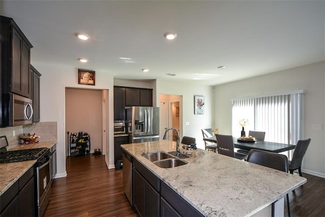 kitchen with appliances with stainless steel finishes, an island with sink, sink, dark hardwood / wood-style flooring, and light stone countertops