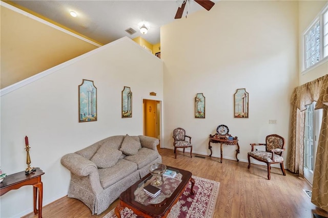 living area featuring visible vents, baseboards, a towering ceiling, light wood-style flooring, and ceiling fan