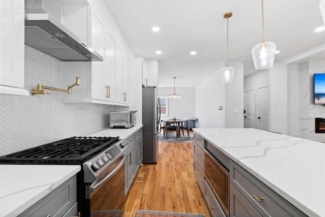 kitchen with gray cabinetry, light wood finished floors, wall chimney range hood, appliances with stainless steel finishes, and tasteful backsplash