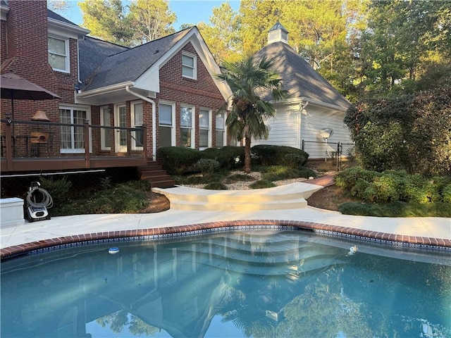 rear view of property featuring an outdoor pool, brick siding, a deck, and fence