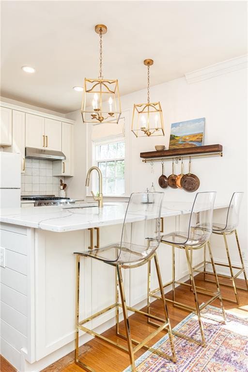 kitchen with light wood-style flooring, tasteful backsplash, an inviting chandelier, light stone counters, and under cabinet range hood