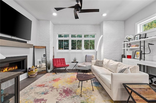 living room with a ceiling fan, a warm lit fireplace, wood finished floors, recessed lighting, and brick wall