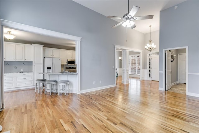 unfurnished living room with baseboards, light wood-style flooring, a high ceiling, ornate columns, and ceiling fan with notable chandelier