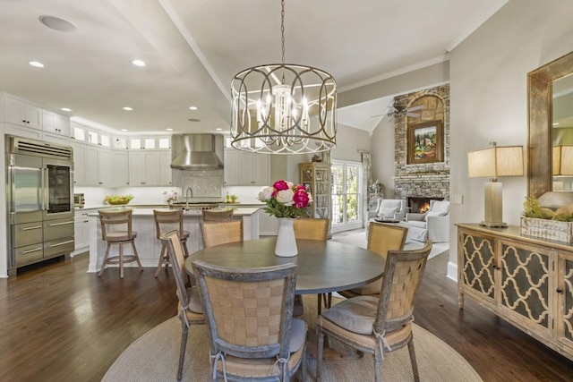 dining space featuring dark wood finished floors, a stone fireplace, ornamental molding, and ceiling fan with notable chandelier