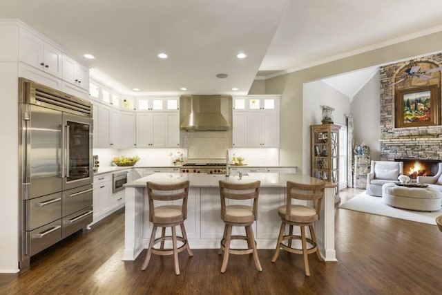 kitchen with wall chimney range hood, built in appliances, light countertops, white cabinets, and dark wood-style floors