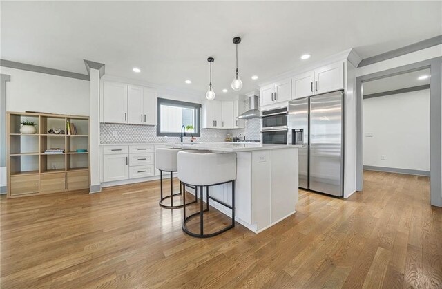 kitchen with wall chimney range hood, appliances with stainless steel finishes, white cabinets, a kitchen island, and decorative light fixtures