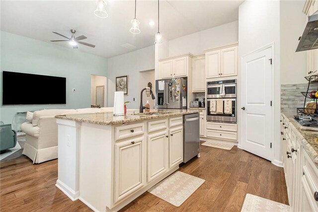 kitchen with cream cabinets, ventilation hood, an island with sink, decorative light fixtures, and stainless steel appliances