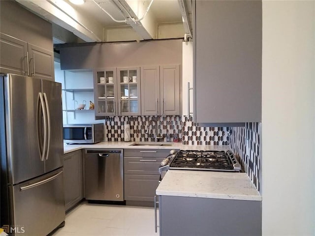 kitchen featuring gray cabinets, backsplash, appliances with stainless steel finishes, glass insert cabinets, and a sink