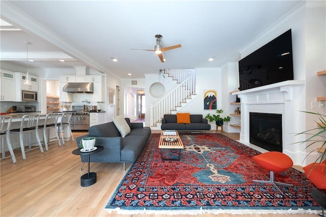 living room with light wood finished floors, crown molding, a fireplace, ceiling fan, and stairs