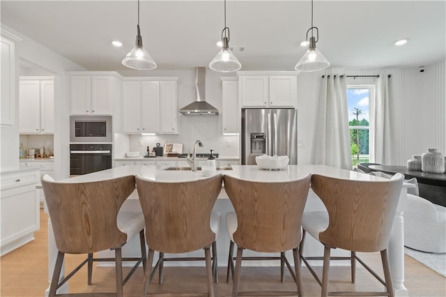 kitchen with wall chimney range hood, oven, a center island with sink, white cabinetry, and stainless steel fridge