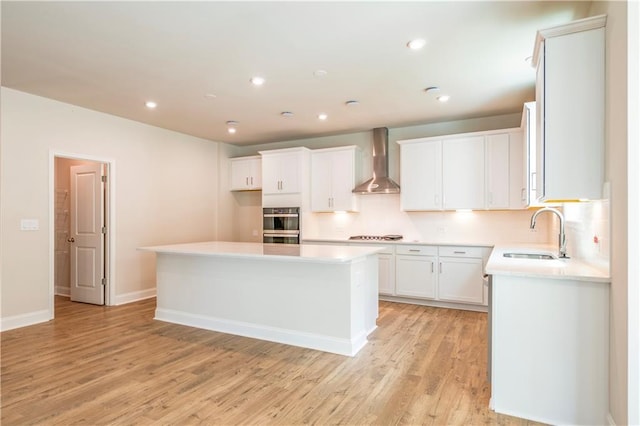 kitchen featuring sink, white cabinets, light wood-type flooring, wall chimney range hood, and a kitchen island