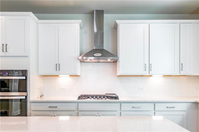 kitchen featuring stainless steel gas stovetop, tasteful backsplash, multiple ovens, wall chimney range hood, and white cabinets