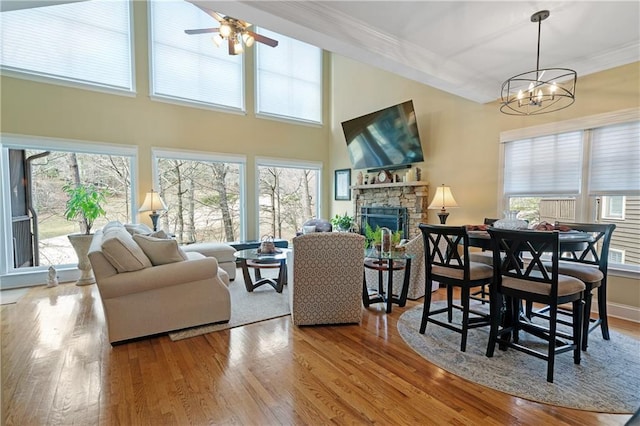 living room with a stone fireplace, ceiling fan with notable chandelier, wood finished floors, a towering ceiling, and crown molding