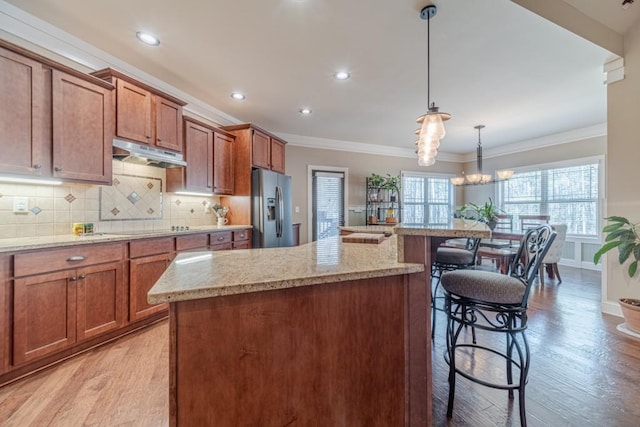 kitchen featuring under cabinet range hood, brown cabinetry, ornamental molding, and stainless steel fridge with ice dispenser