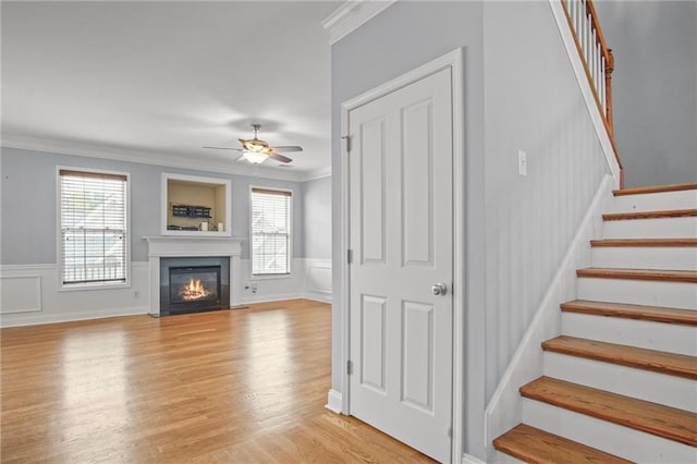 unfurnished living room featuring light wood-type flooring, a fireplace with flush hearth, ornamental molding, a ceiling fan, and stairs