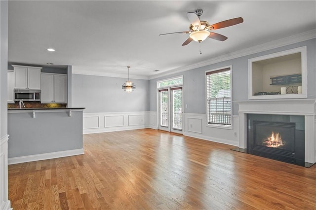 unfurnished living room with ornamental molding, ceiling fan, wainscoting, a glass covered fireplace, and light wood-type flooring