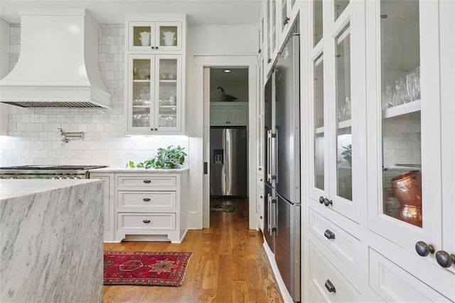 kitchen with dark wood-type flooring, white cabinetry, custom range hood, stainless steel appliances, and backsplash