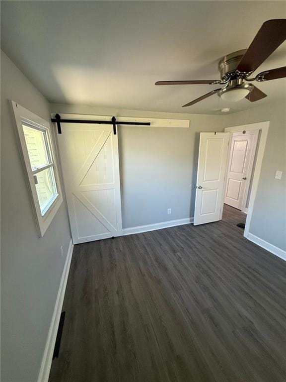 unfurnished bedroom with a barn door, baseboards, dark wood-style floors, and a ceiling fan