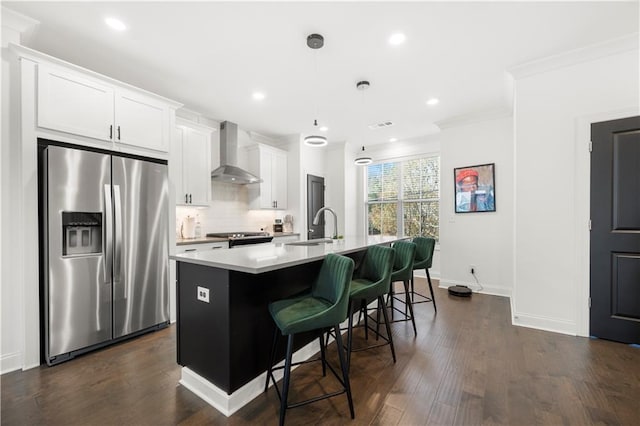 kitchen with a breakfast bar, stainless steel appliances, wall chimney range hood, white cabinetry, and an island with sink