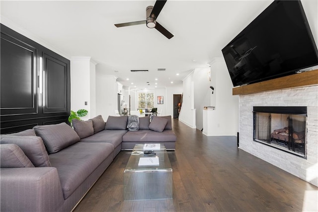 living room with crown molding, ceiling fan, a stone fireplace, and dark wood-type flooring