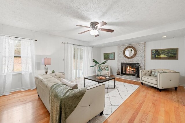 living room with a brick fireplace, ceiling fan, a textured ceiling, and light wood-type flooring