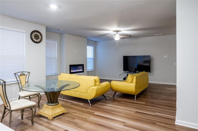 living room with light wood-style flooring, visible vents, a ceiling fan, baseboards, and a glass covered fireplace