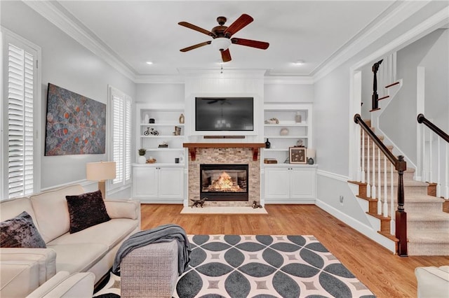 living area featuring light wood finished floors, ceiling fan, a fireplace with flush hearth, crown molding, and stairs