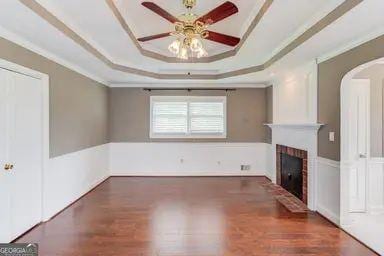 unfurnished living room with ceiling fan, a fireplace, arched walkways, wood finished floors, and a tray ceiling