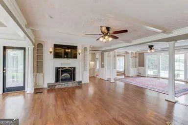 unfurnished living room featuring a fireplace with raised hearth, decorative columns, ceiling fan, and wood finished floors
