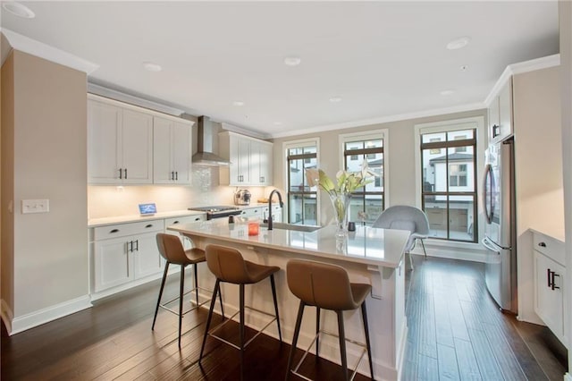 kitchen featuring wall chimney range hood, sink, stainless steel fridge, white cabinets, and a center island with sink