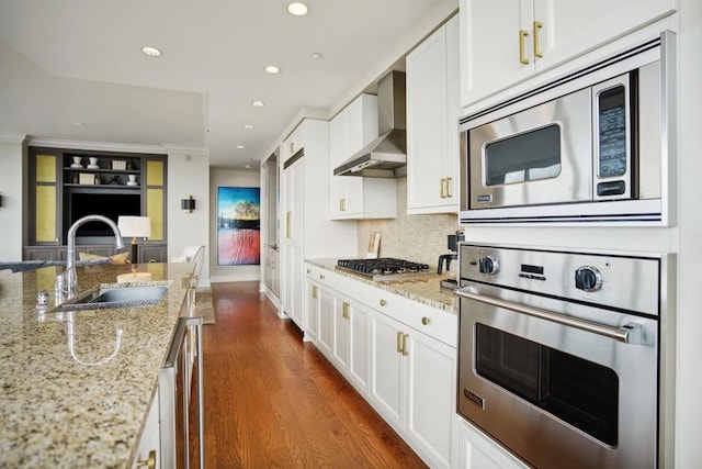 kitchen featuring light stone counters, wall chimney exhaust hood, stainless steel appliances, sink, and white cabinetry