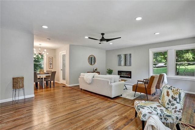 living room featuring recessed lighting, a glass covered fireplace, wood finished floors, baseboards, and ceiling fan with notable chandelier